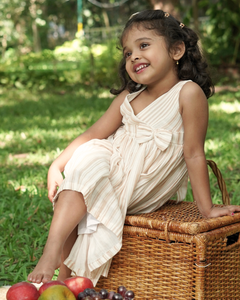 Young girl in the Faye Striped Beige Dress sitting on a wicker basket outdoors with greenery in the background