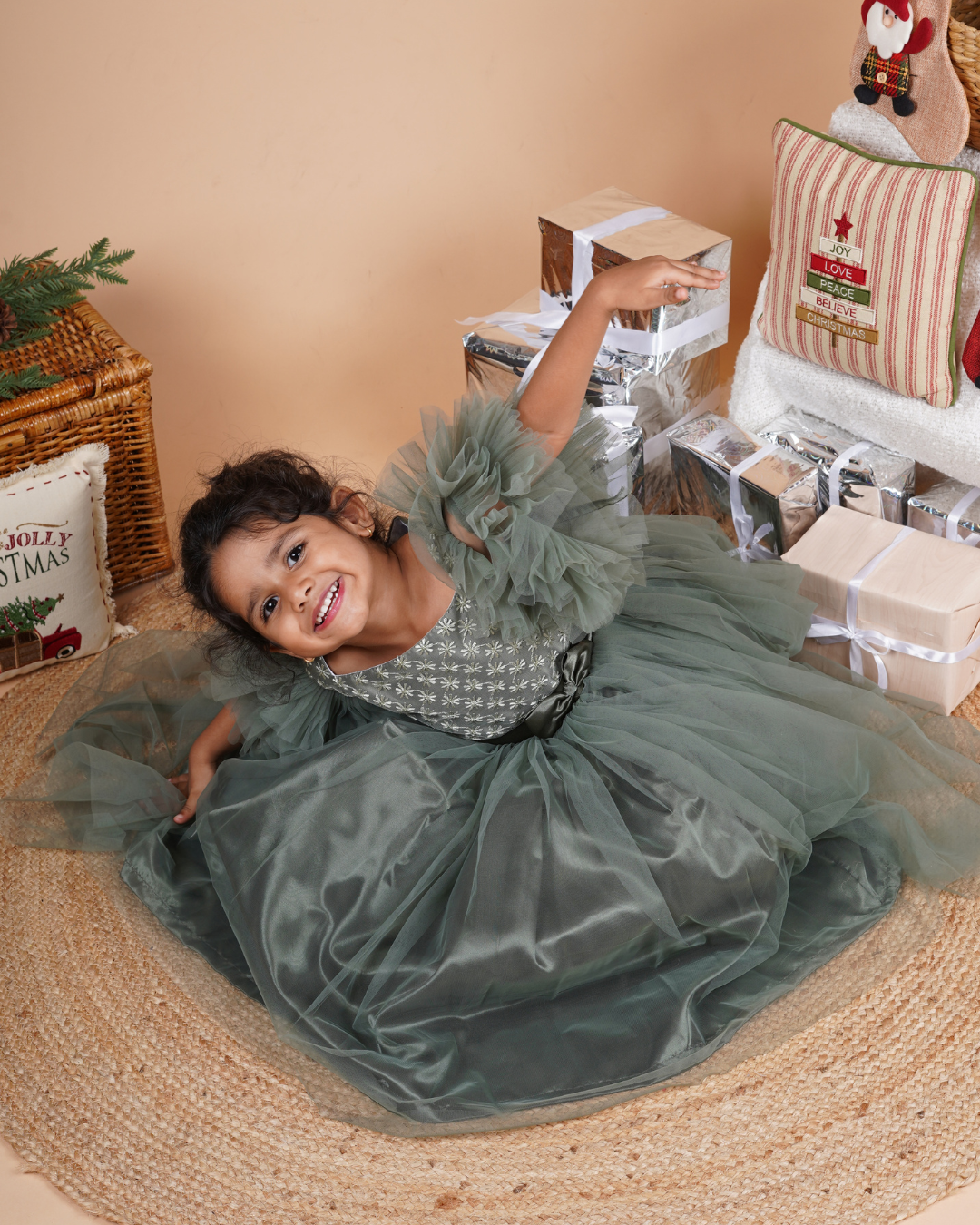 Young girl in the Faye Green Tiered Sleeve Gown sitting on a woven mat with Christmas decorations in the background.