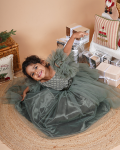Young girl in the Faye Green Tiered Sleeve Gown sitting on a woven mat with Christmas decorations in the background.
