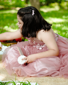 Young girl in the Faye Mauve One Shoulder Gown sitting on grass with a book and flowers.