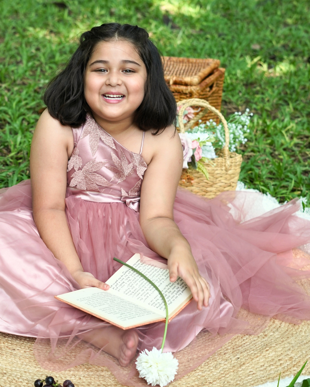 Young girl in the Faye Mauve One Shoulder Gown sitting on grass with a book and flowers.