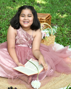 Young girl in the Faye Mauve One Shoulder Gown sitting on grass with a book and flowers.