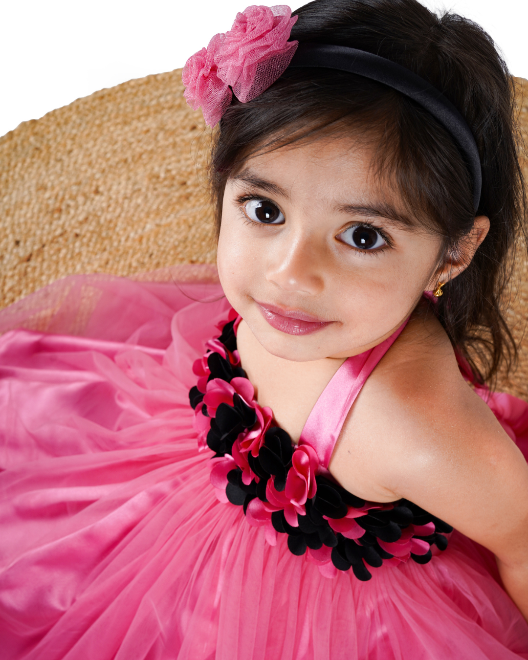 Young girl in the Faye Onion Pink Tutu Dress a pink flower hairband in her hair, sitting on a woven mat.