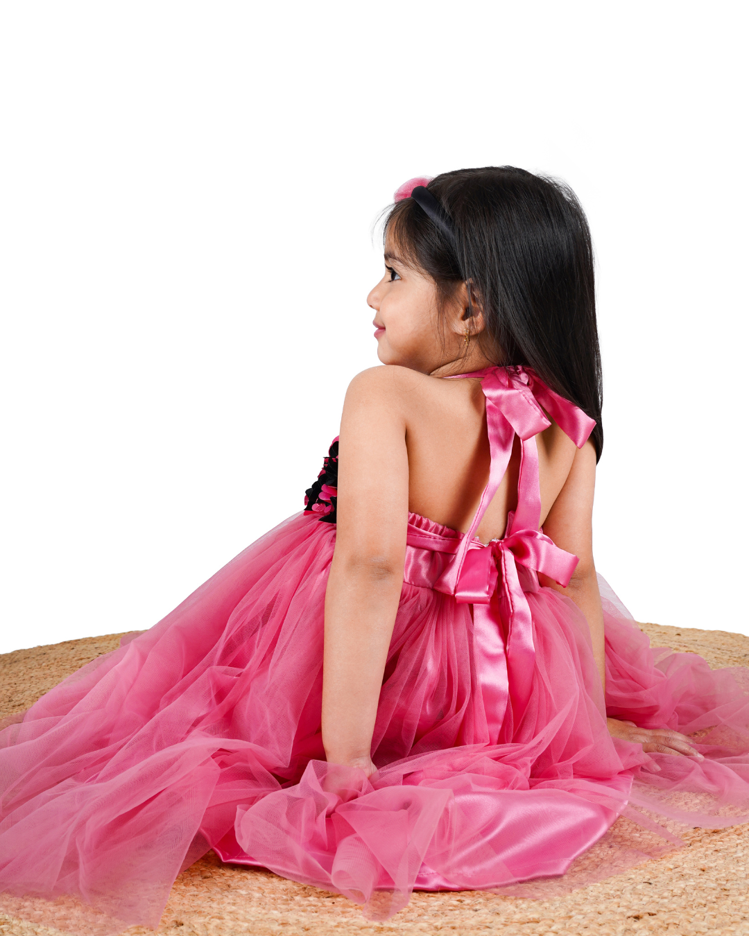 Young girl in the Faye Onion Pink Tutu Dress a pink flower hairband in her hair, sitting on a woven mat.