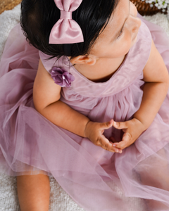 Toddler in the Faye Taupe Swing Dress with a bow and flower, sitting on a textured surface.