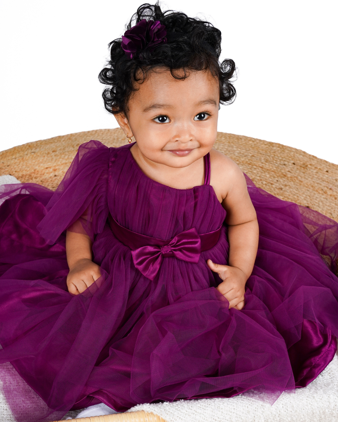 Baby in the Faye Wine One Shoulder Dress with a bow sitting on a woven surface.